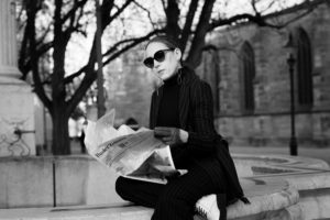 Woman in black with sunglasses sitting on fountain reading newspaper